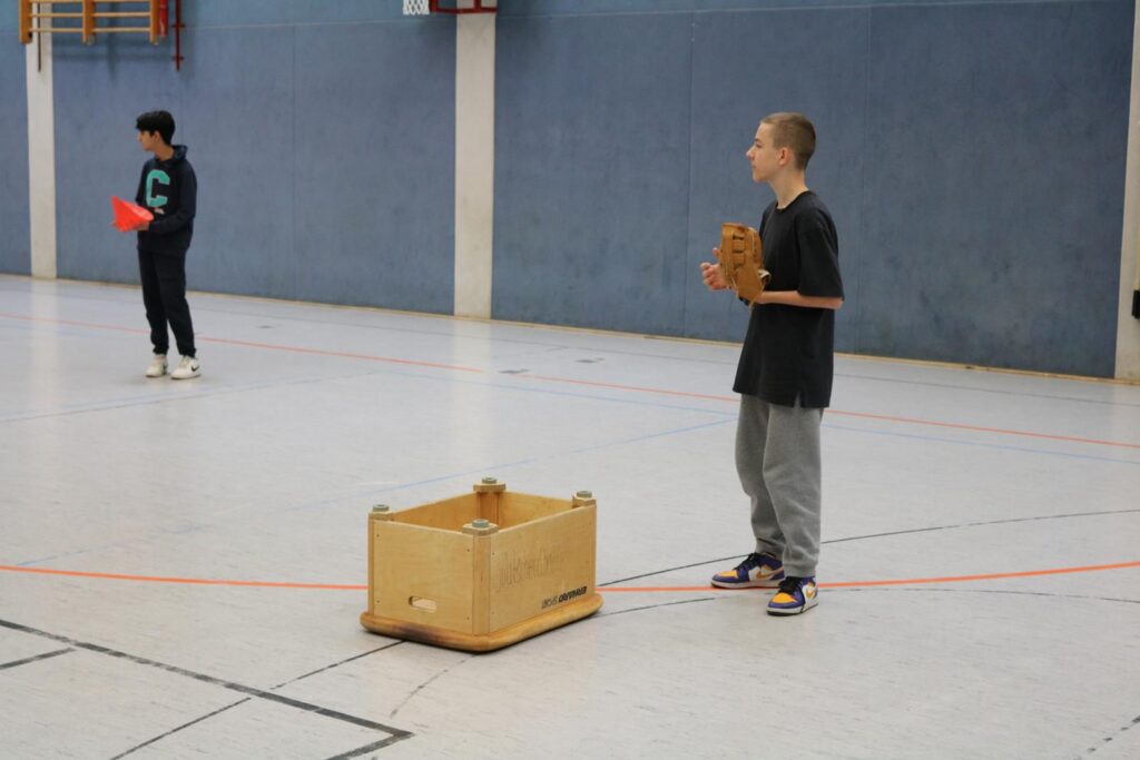 Zwei Kinder in einer Sporthalle, eines mit Handschuh und Holzklotz.