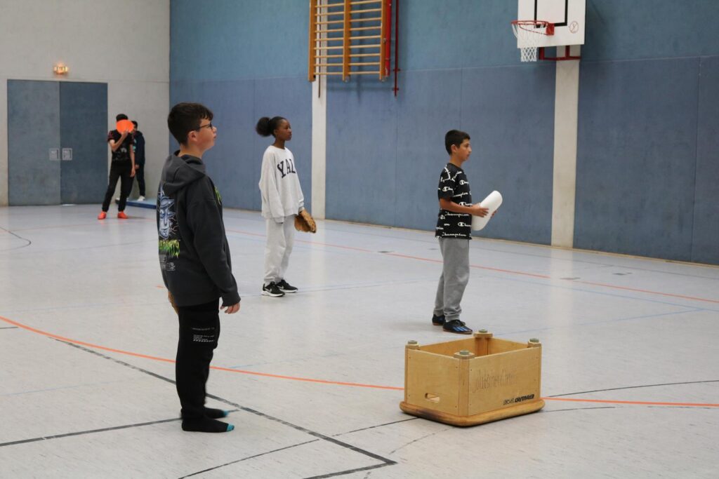 Kinder stehen in einer Sporthalle, einige halten Gegenstände in der Hand.