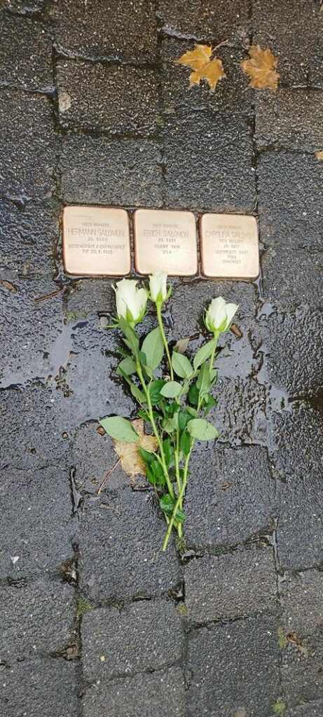 Drei Stolpersteine mit weißen Rosen auf einem Gehweg.