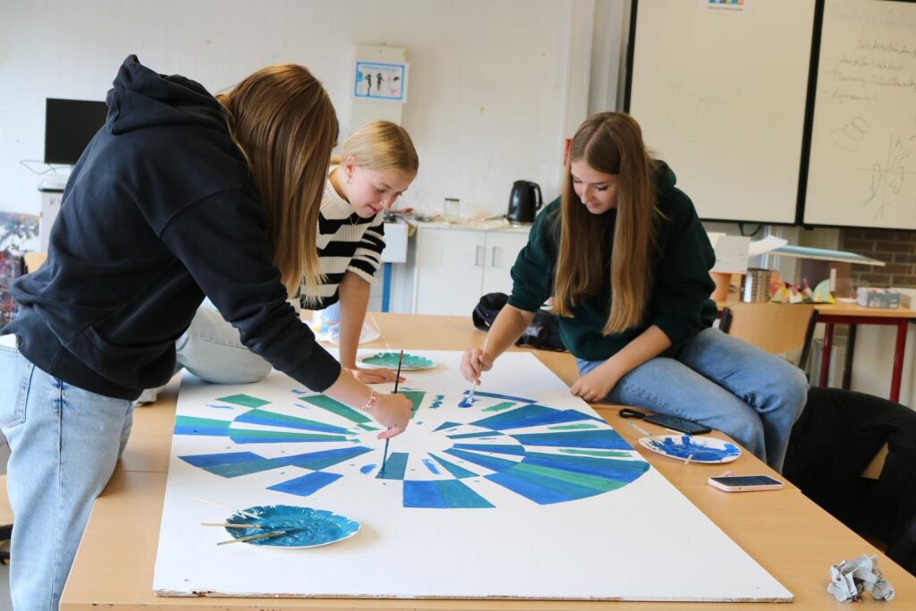 Drei Schüler:innen arbeiten an einem Kunstprojekt mit Farben auf einem großen Plakat.