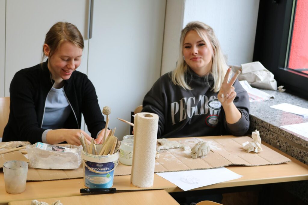Zwei Schülerinnen arbeiten an Tonfiguren und lächeln in einem Kunstraum.