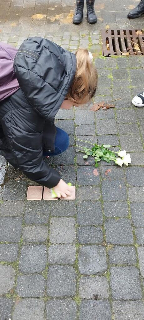 Eine Person kniet sich auf den Gehweg und reinigt einen Gedenkstein. Blumen liegen daneben.