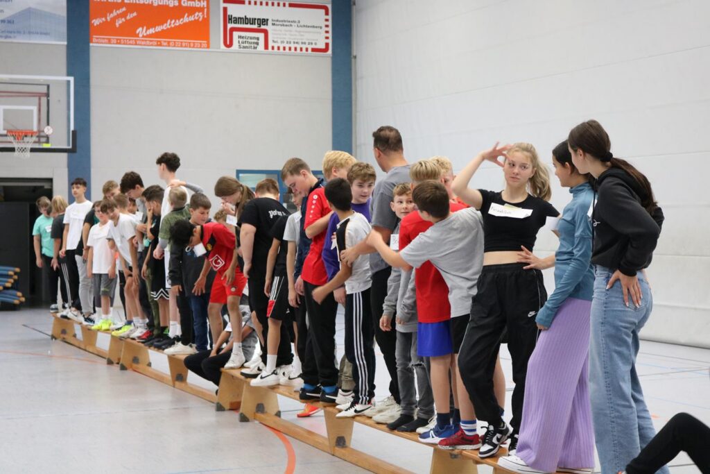 Schüler stehen in einer Reihe auf einem langen Balancebalken in der Sporthalle.
