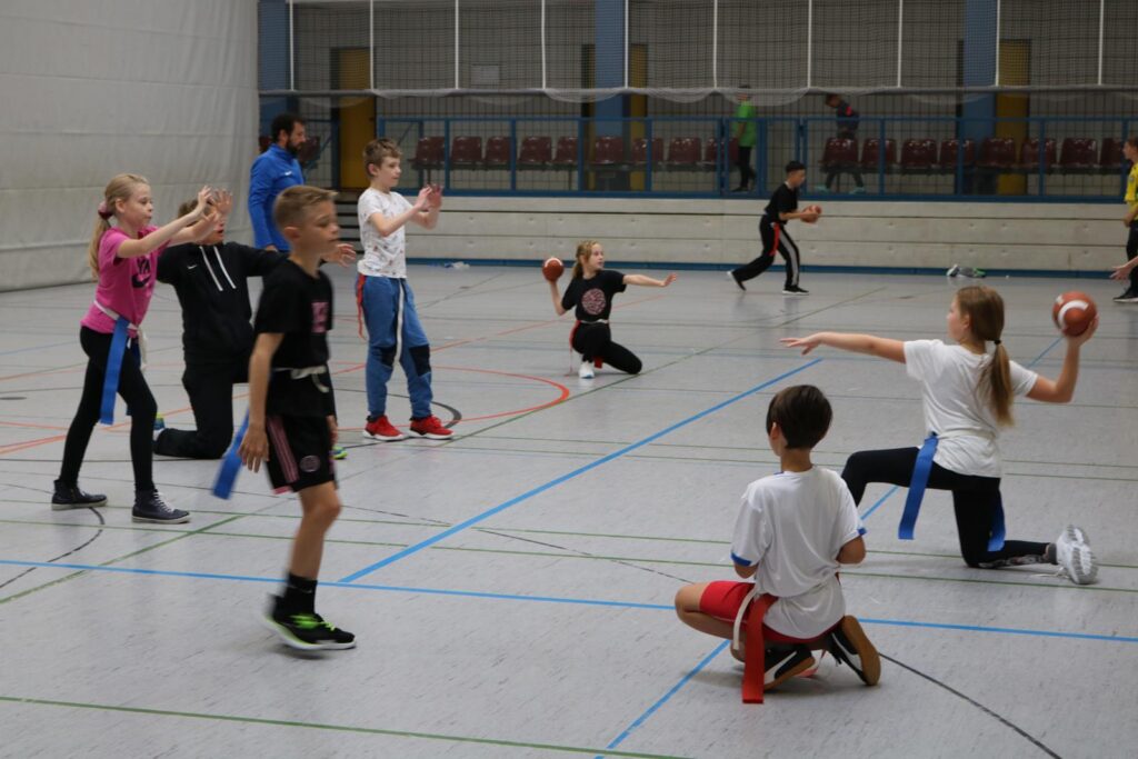 Kinder spielen in einer Sporthalle mit Bällen und blauen Stäben.