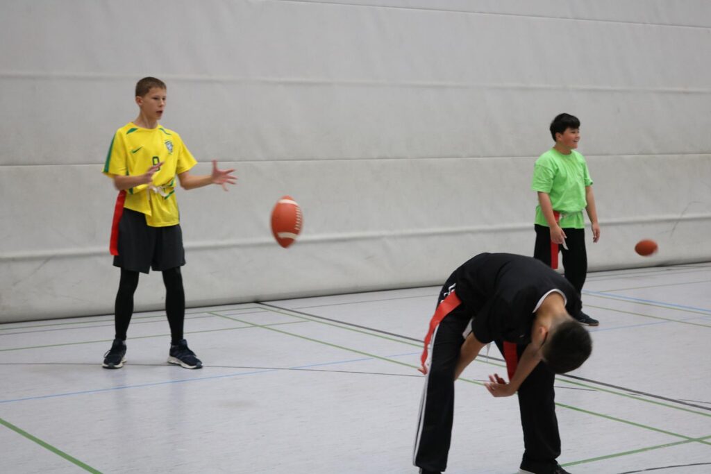 Drei Jugendliche werfen und fangen Footballs in einer Sporthalle.
