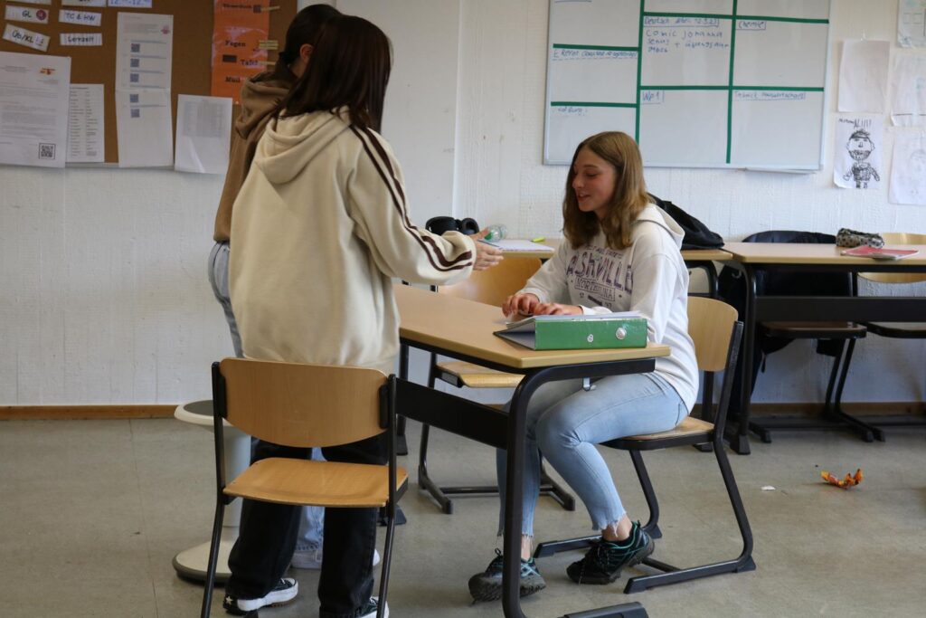 Drei Schülerinnen interagieren in einem Klassenzimmer an einem Tisch.