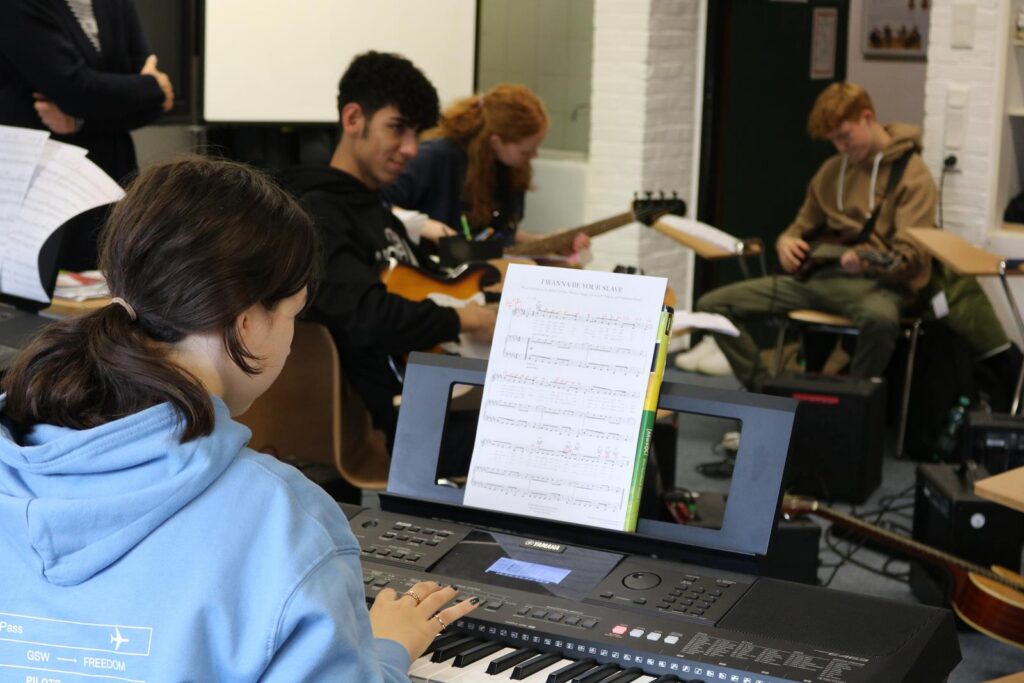Schüler spielen Instrumente in einem Musikraum. Notenblätter liegen auf einem Keyboard.