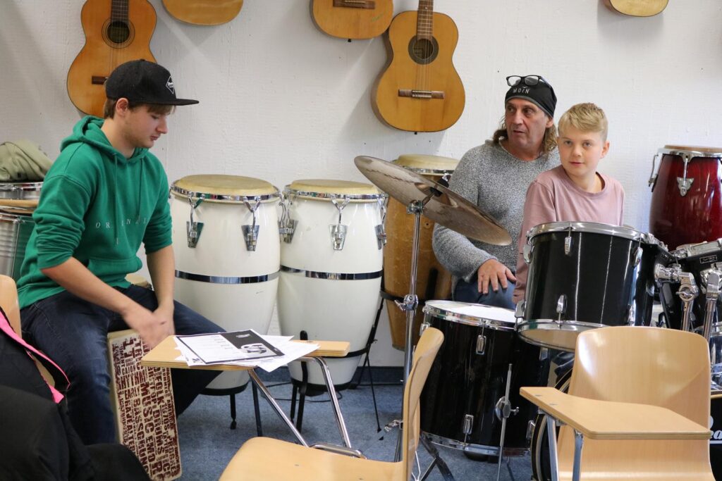 Zwei Schüler und ein Lehrer spielen Schlaginstrumente in einem Musikraum.