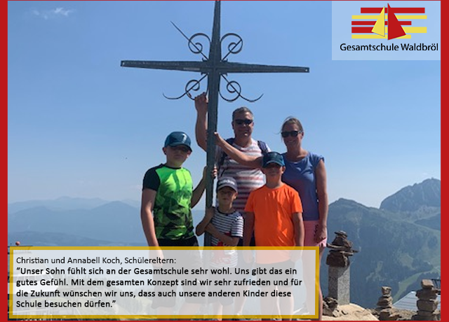 Familie Koch steht mit einem Kreuz auf einem Berggipfel. Hintergrund: Berge und Himmel.