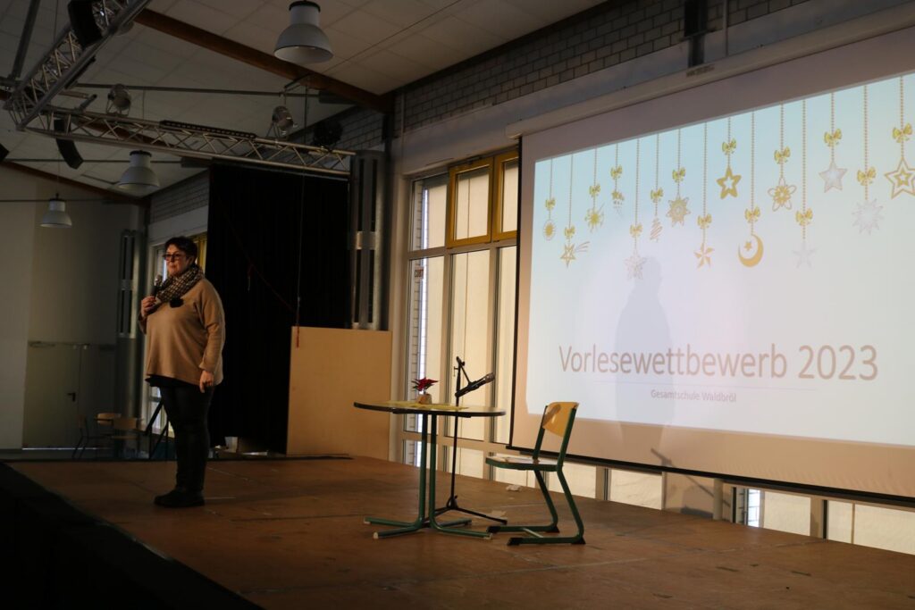 Eine Person steht auf einer Bühne vor einer Präsentation zum Vorlesewettbewerb 2023.