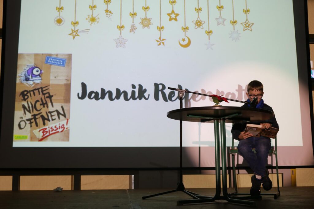 Junger Mann sitzt am Tisch und hält ein Buch, Präsentation im Hintergrund.