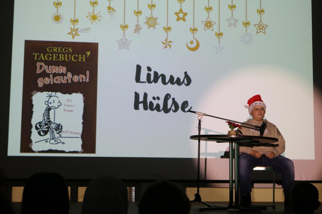 Ein Junge mit einer roten Weihnachtsmütze sitzt am Tisch. Im Hintergrund steht ein Buchcover.
