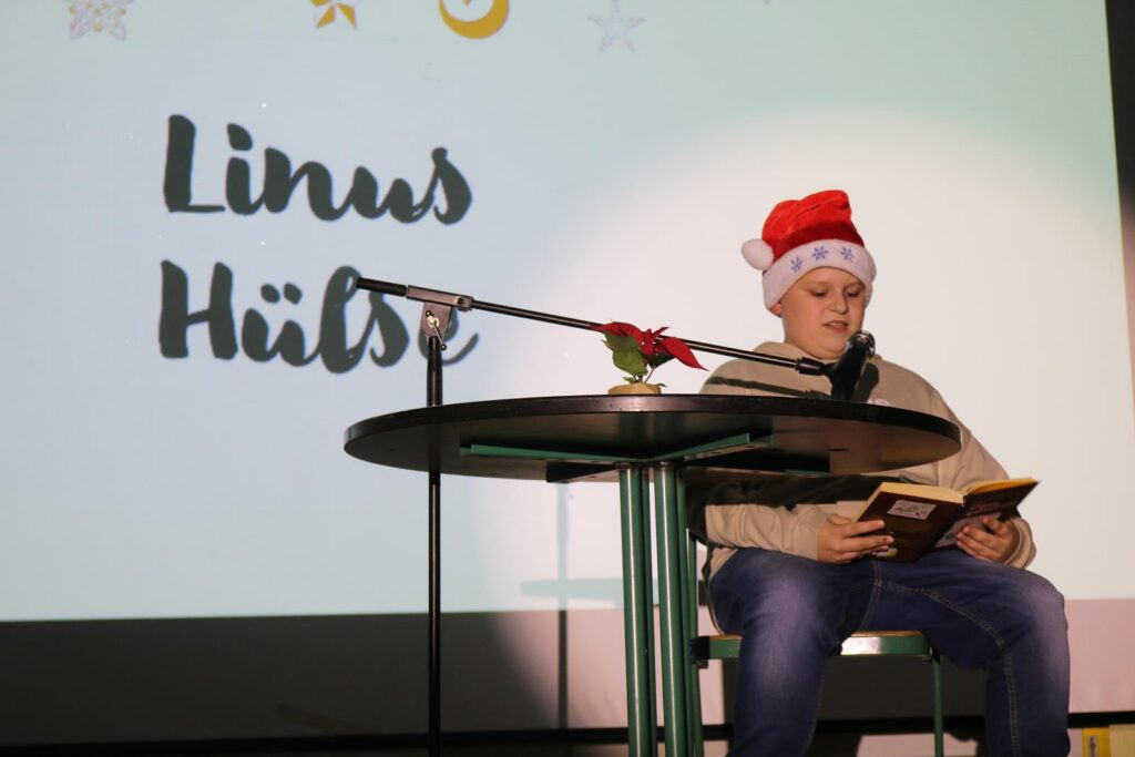 Ein Junge mit Weihnachtsmütze liest an einem Tisch vor Publikum.