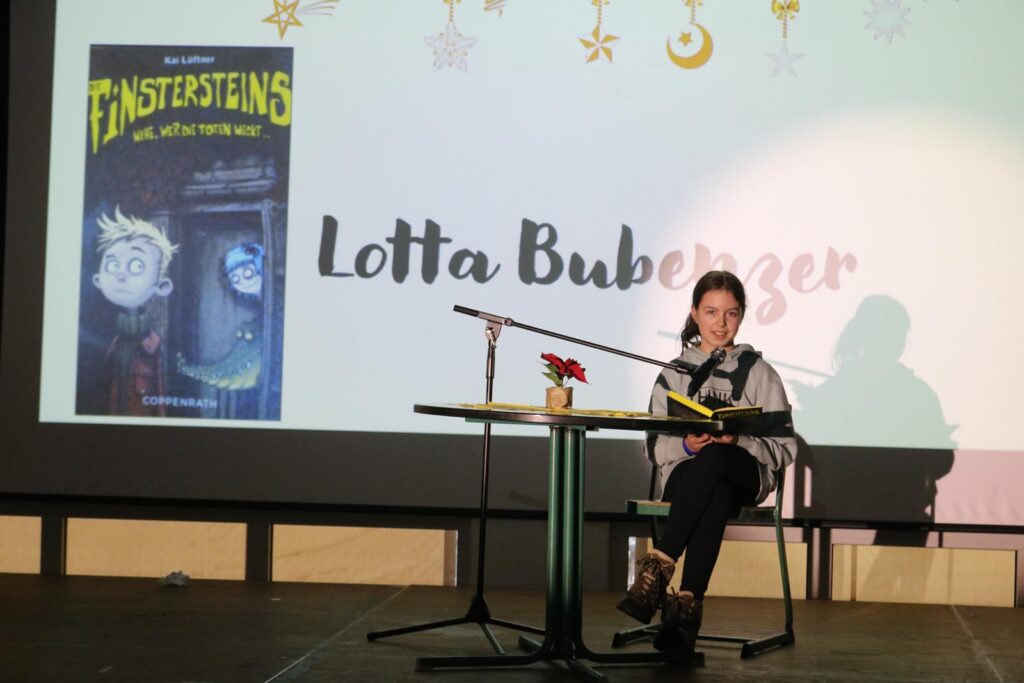 Lotta Bubinger sitzt an einem Tisch und liest aus einem Buch vor.