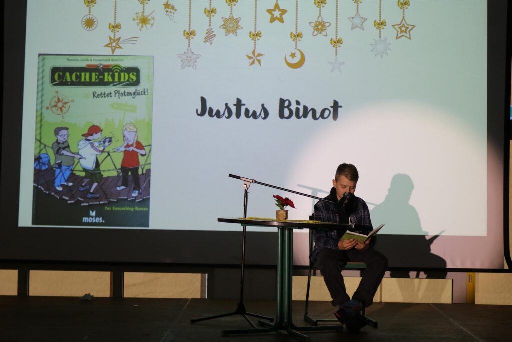 Justus Binot liest aus einem Buch vor, im Hintergrund eine Leuchttafel mit Buchcover.