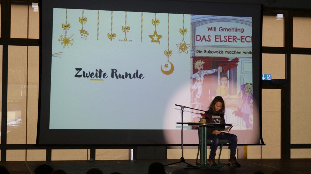 Schülerin sitzt an einem Tisch und präsentiert auf der Bühne. Bildschirm zeigt "Zweite Runde".