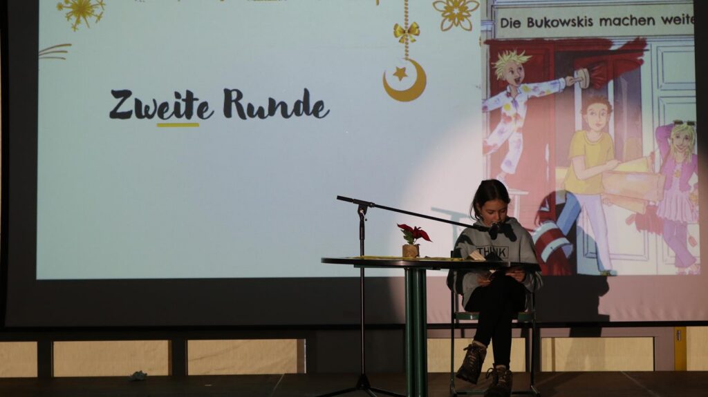 Schülerin sitzt während einer Lesung an einem Tisch auf der Bühne.