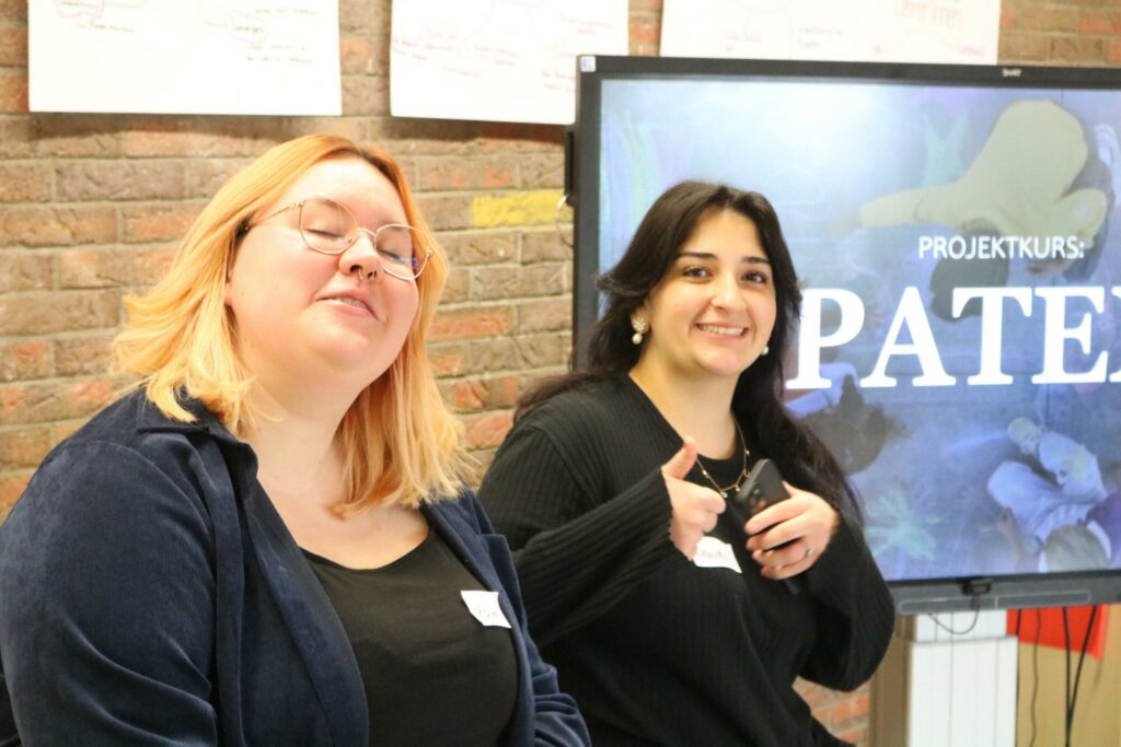 Zwei Personen sitzen lächelnd vor einem Bildschirm mit dem Wort "Paten".