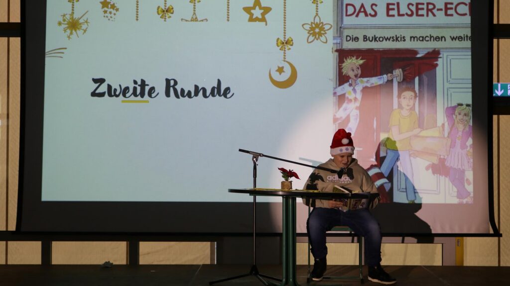 Junge sitzt am Tisch, trägt eine Weihnachtsmütze, liest vor einer Leinwand vor.