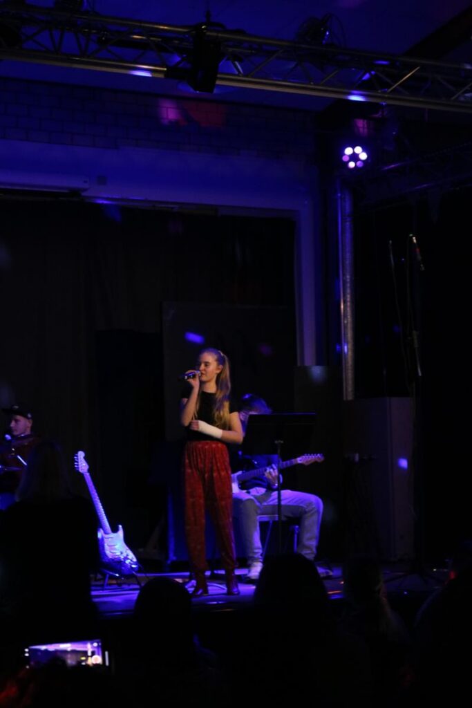 Schülerin singt auf der Bühne, begleitet von zwei Musikern im Hintergrund.