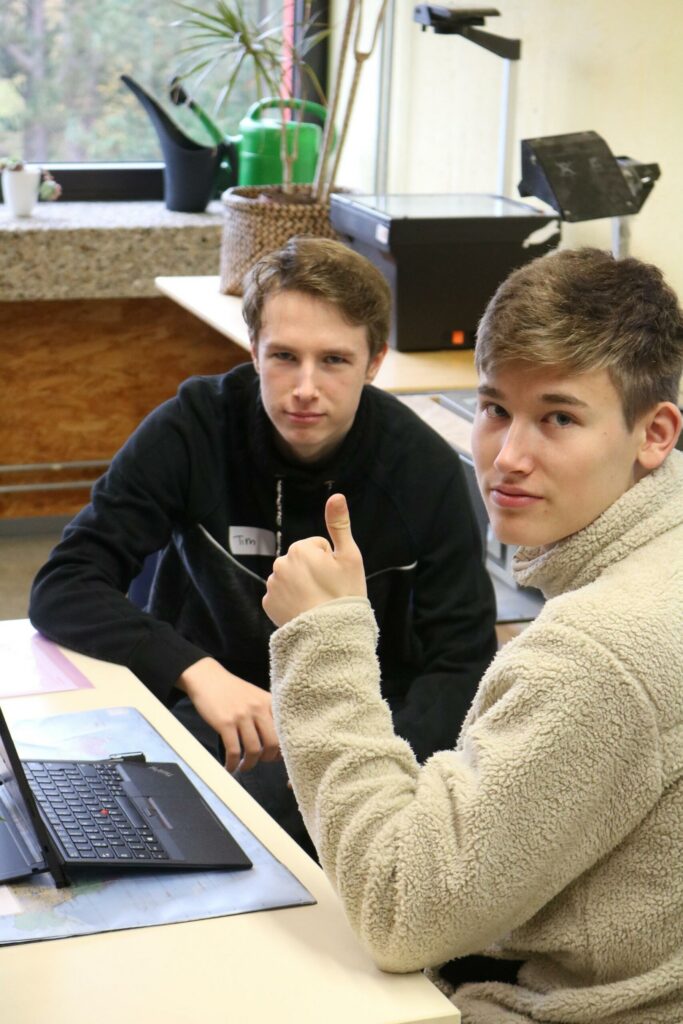 Zwei Schüler sitzen an einem Tisch mit einem Laptop und schauen in die Kamera.