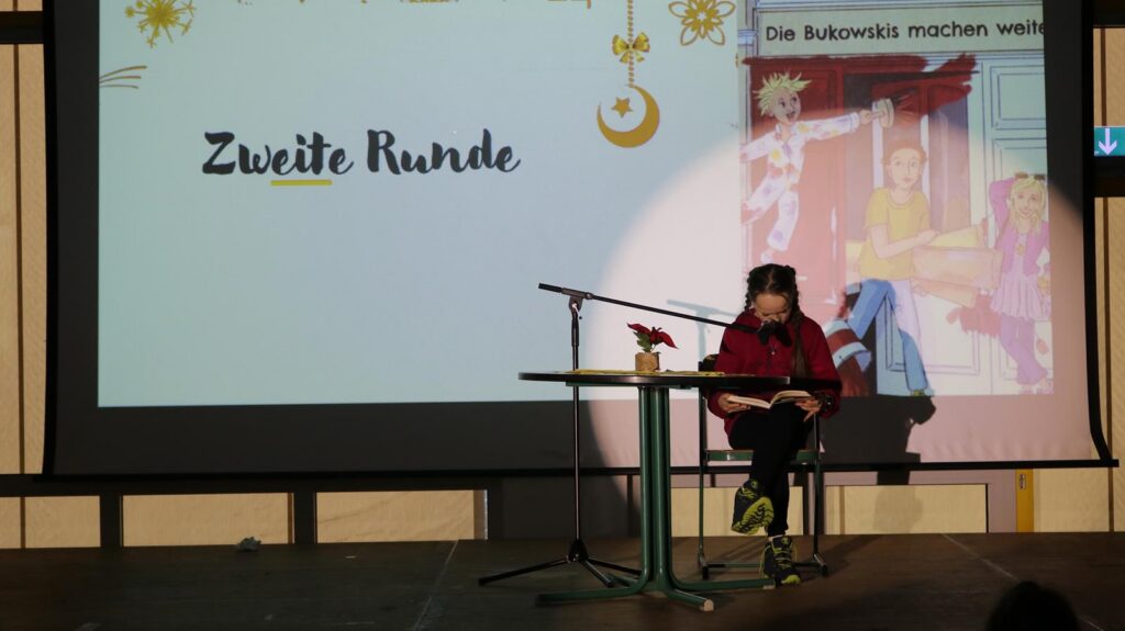 Schüler sitzt am Tisch und liest auf einer Bühne vor Publikum. Hintergrund mit Präsentation.