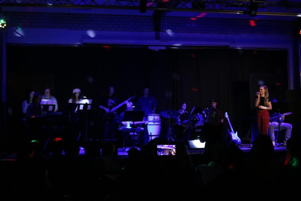 Musikaufführung mit Band und Sängerin auf einer Bühne, bunte Lichter im Hintergrund.