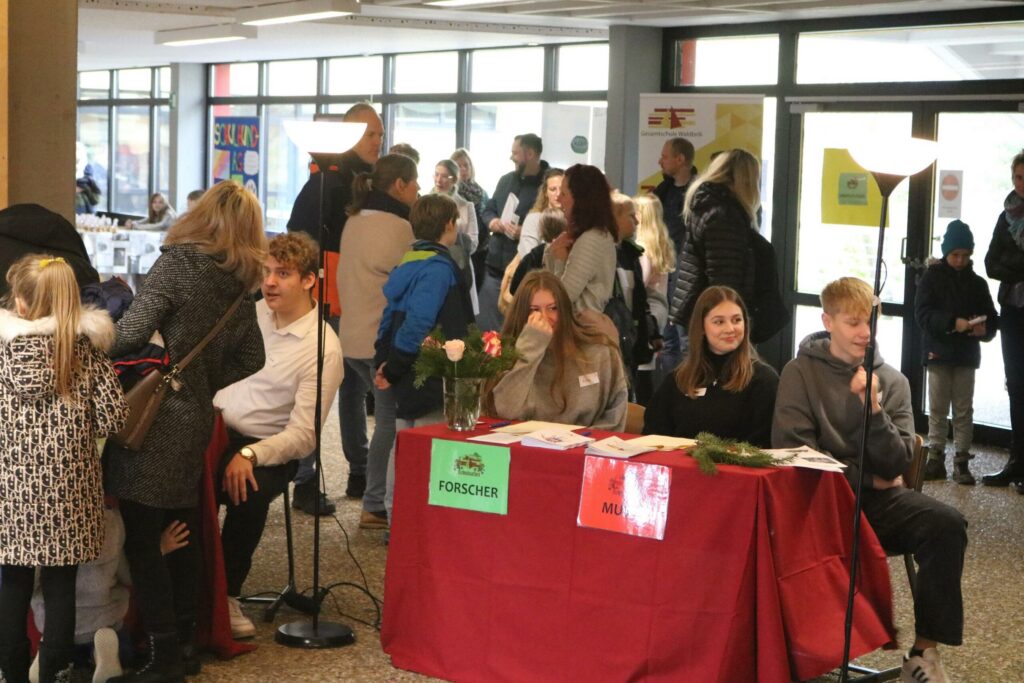 Menschen bei einer Veranstaltung im Schulgebäude, Stände und Interaktionen sichtbar.