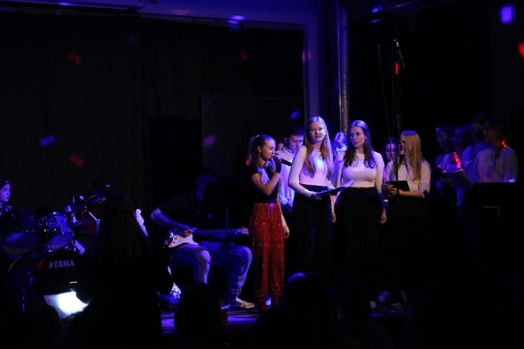 Schülerinnen singen auf einer Bühne, Band im Hintergrund, bunte Lichter.