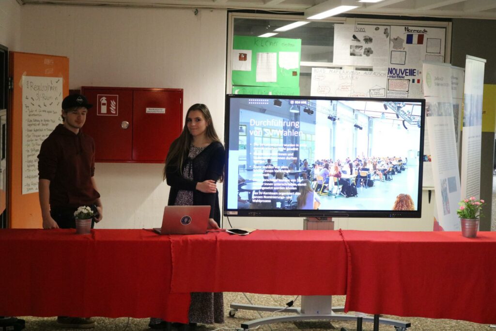 Zwei Personen stehen an einem Tisch mit Laptop vor einer Präsentationswand.