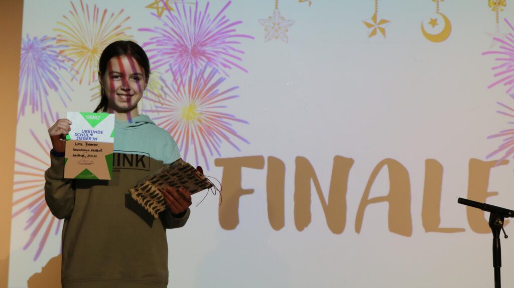 Schülerin hält eine Auszeichnung vor festlicher Kulisse mit dem Wort "FINALE".