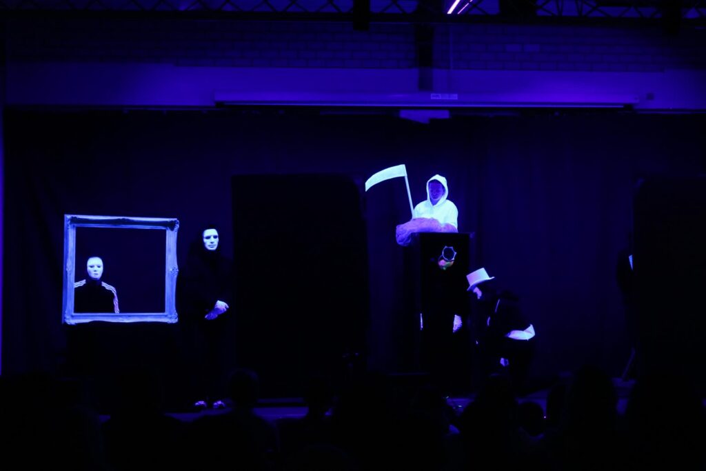 Szenenbild einer Theateraufführung mit schwarzem Hintergrund und leuchtenden Figuren.
