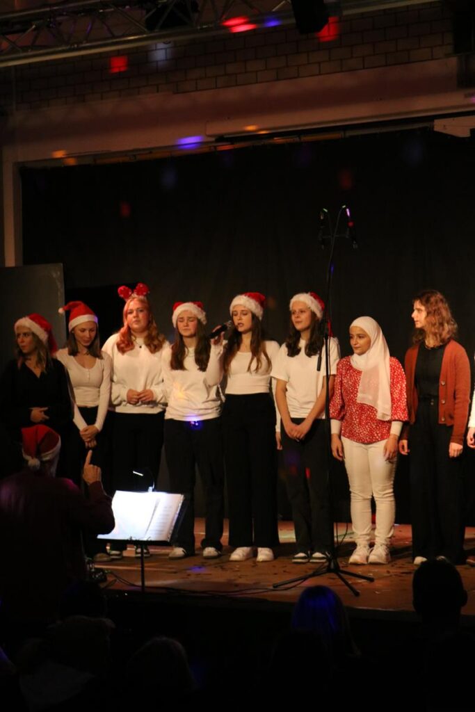 Schülerinnen in Weihnachtsmützen stehen auf der Bühne und singen.