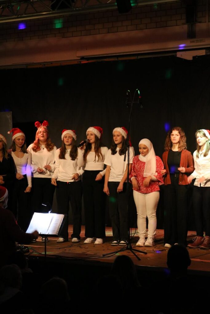 Schülerinnen singen auf einer Bühne, tragen Weihnachtsmützen und Festkleidung.
