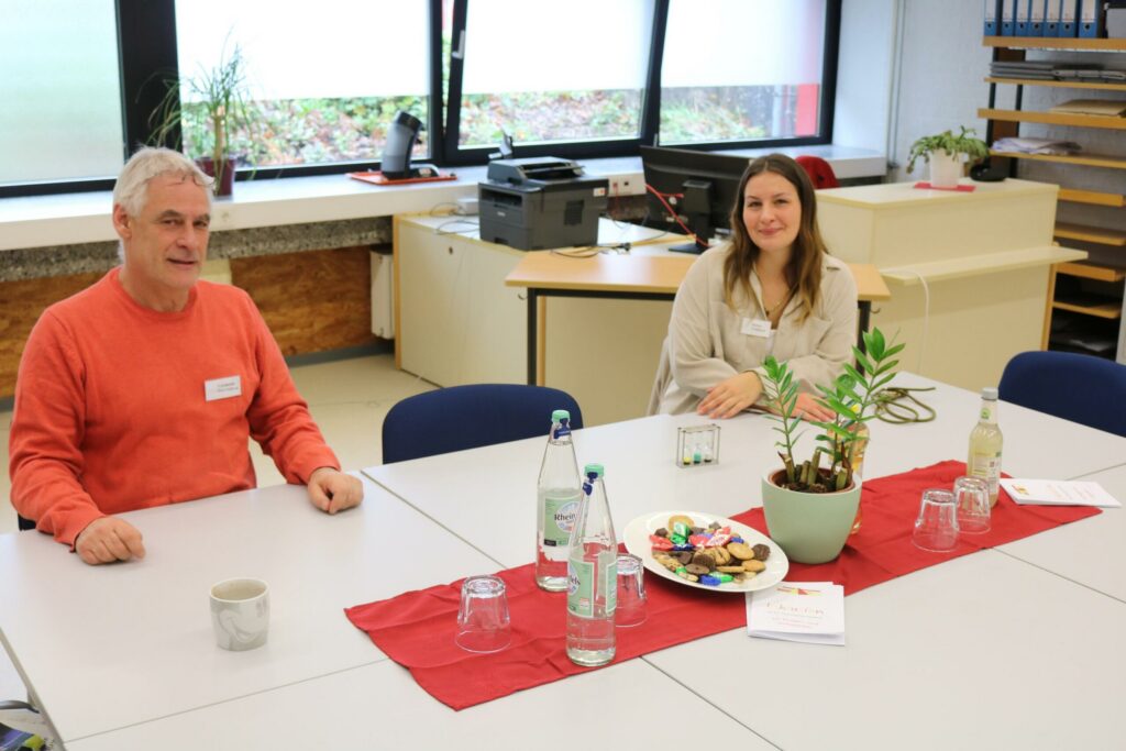 Zwei Personen sitzen an einem Tisch mit Getränke, Keksen und einer Pflanze.