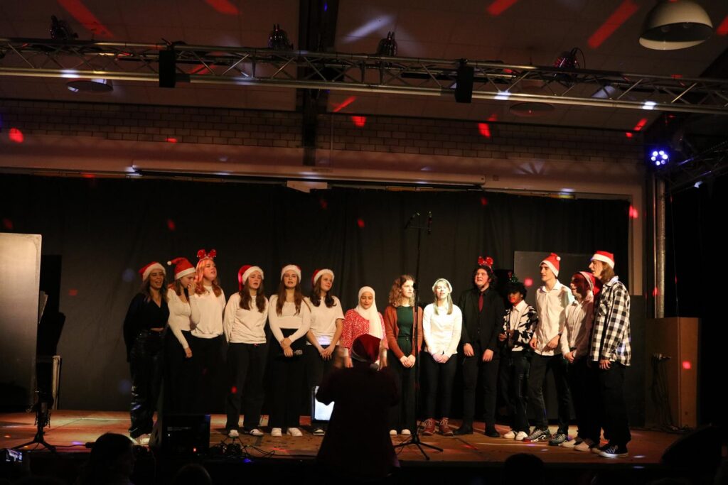 Schulkinder in Weihnachtsmützen singen auf einer Bühne.