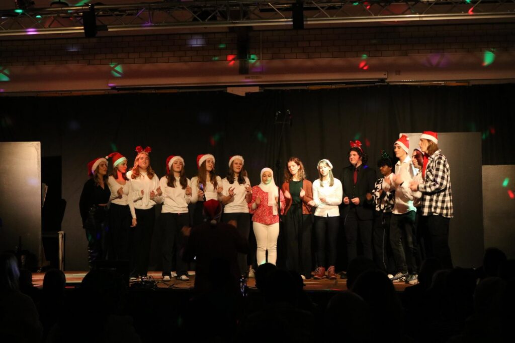 Schülerinnen und Schüler in Weihnachtsmützen stehen auf der Bühne und singen.