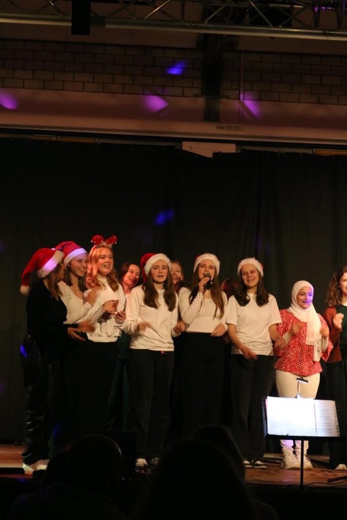 Schüler in Weihnachtsmützen singen auf einer Bühne.