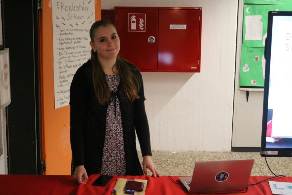 Schülerin steht an einem Tisch mit Laptop und Präsentationsmaterialien.