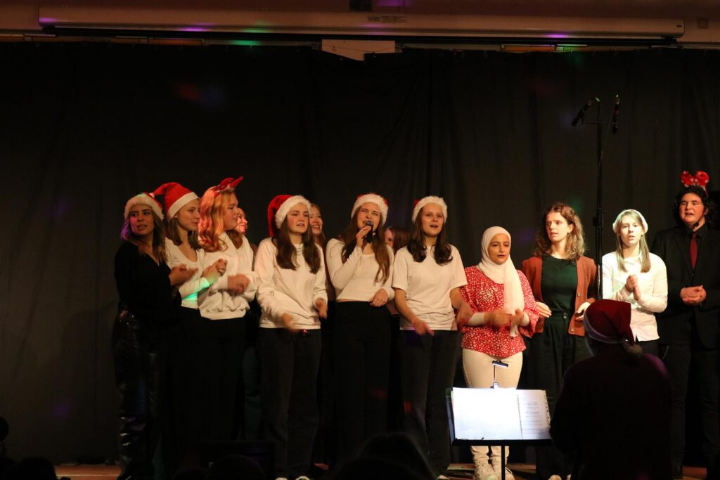 Schüler singen auf einer Bühne, tragen Weihnachtsmützen und festliche Kleidung.