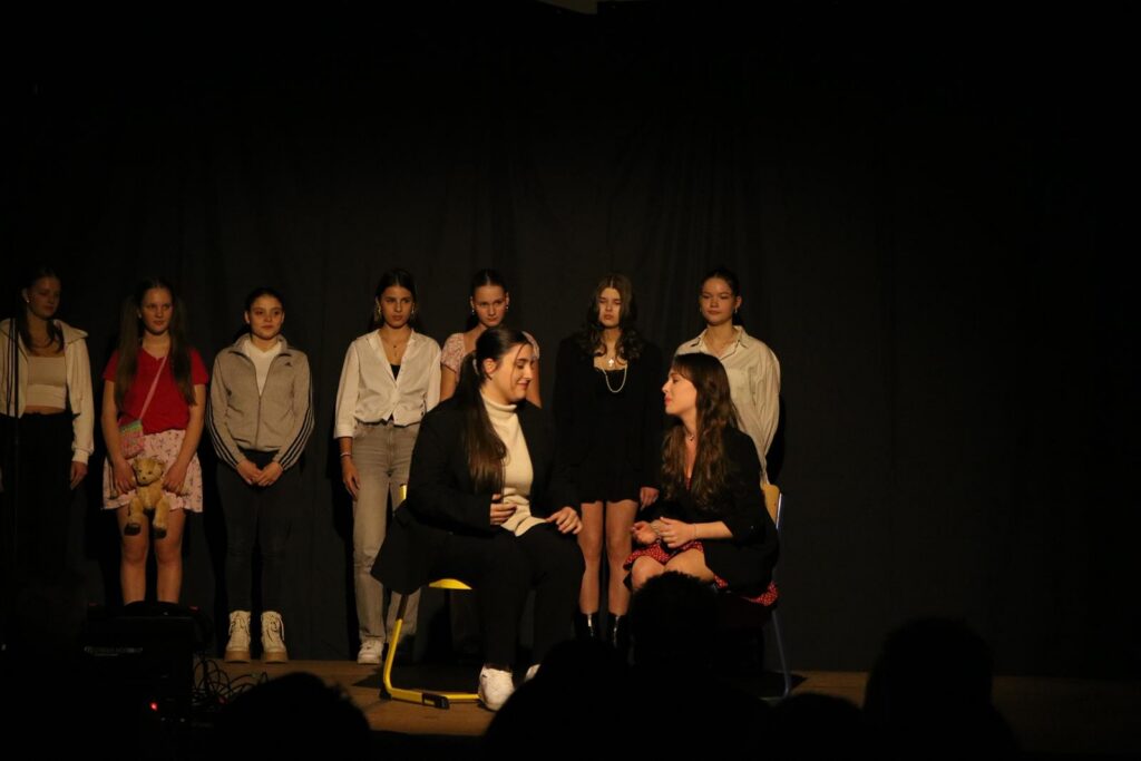 Theateraufführung mit mehreren Schauspielern auf der Bühne, zwei im Vordergrund im Dialog.
