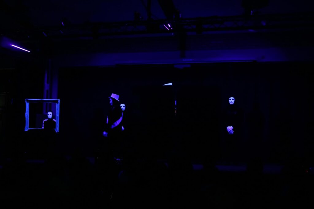 Szenenbild mit mehreren Personen in dunkler Kleidung vor schwarzem Hintergrund.