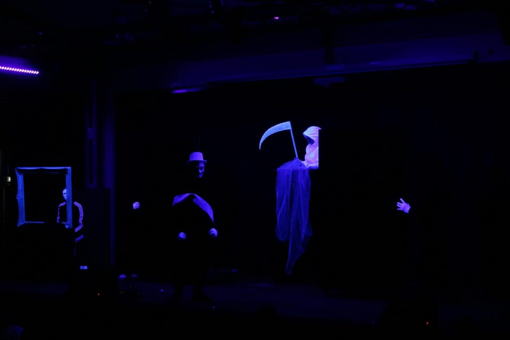 Dunkle Bühnenaufnahme mit Licht- und Schattenspielen, Figur mit Sense.
