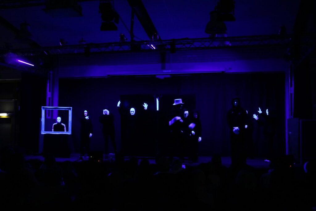 Theateraufführung mit düsterer Beleuchtung und Figuren in Schwarz mit leuchtenden Händen.