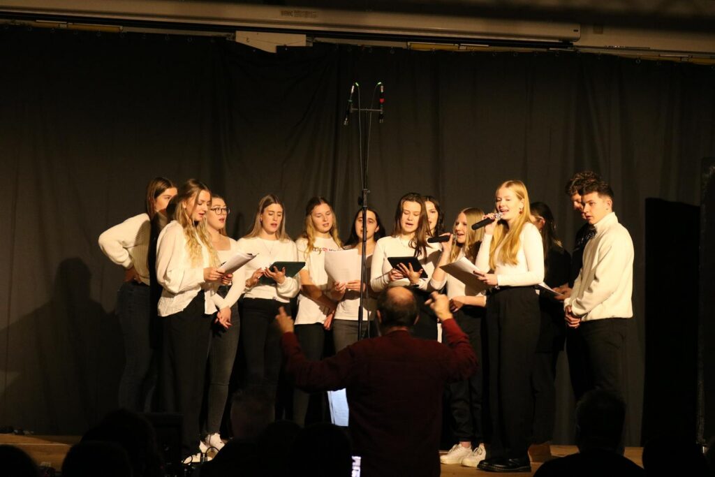 Chor mit Jugendlichen auf der Bühne, sie singen und halten Notenblätter.