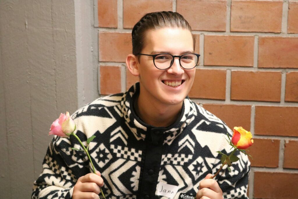 Ein junger Mann hält je eine Blume in jeder Hand und lächelt.