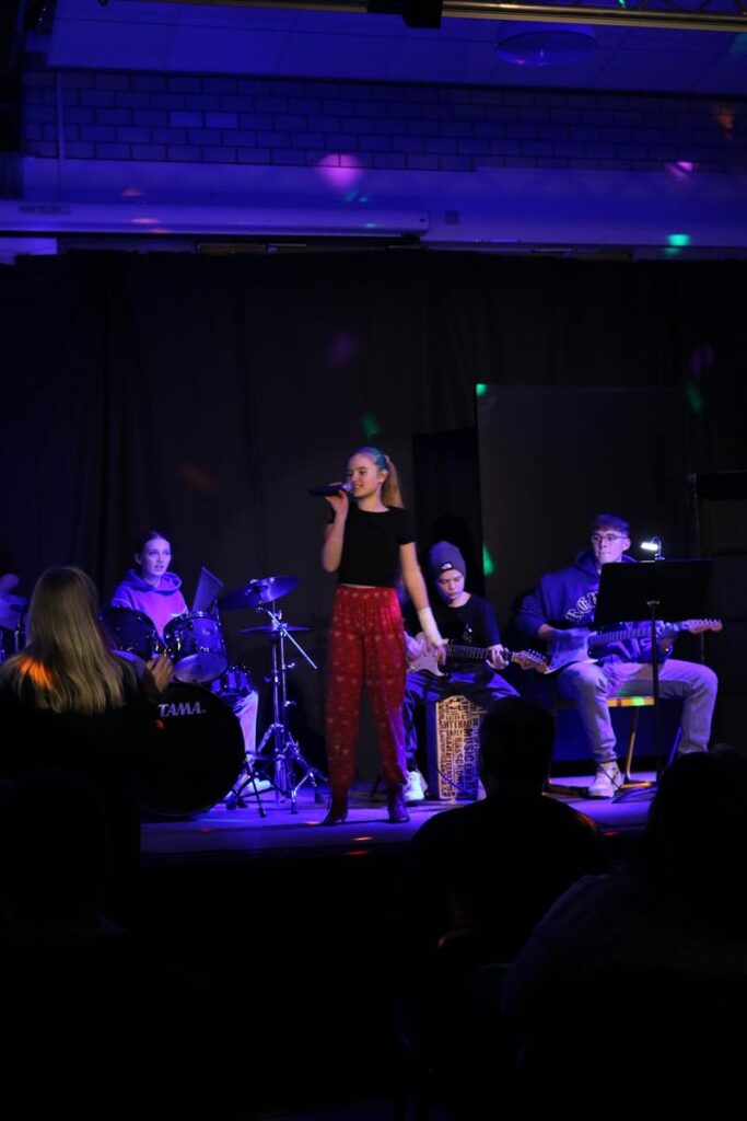 Schülerin singt auf der Bühne, Begleitband im Hintergrund. Beleuchtung in bunten Farben.
