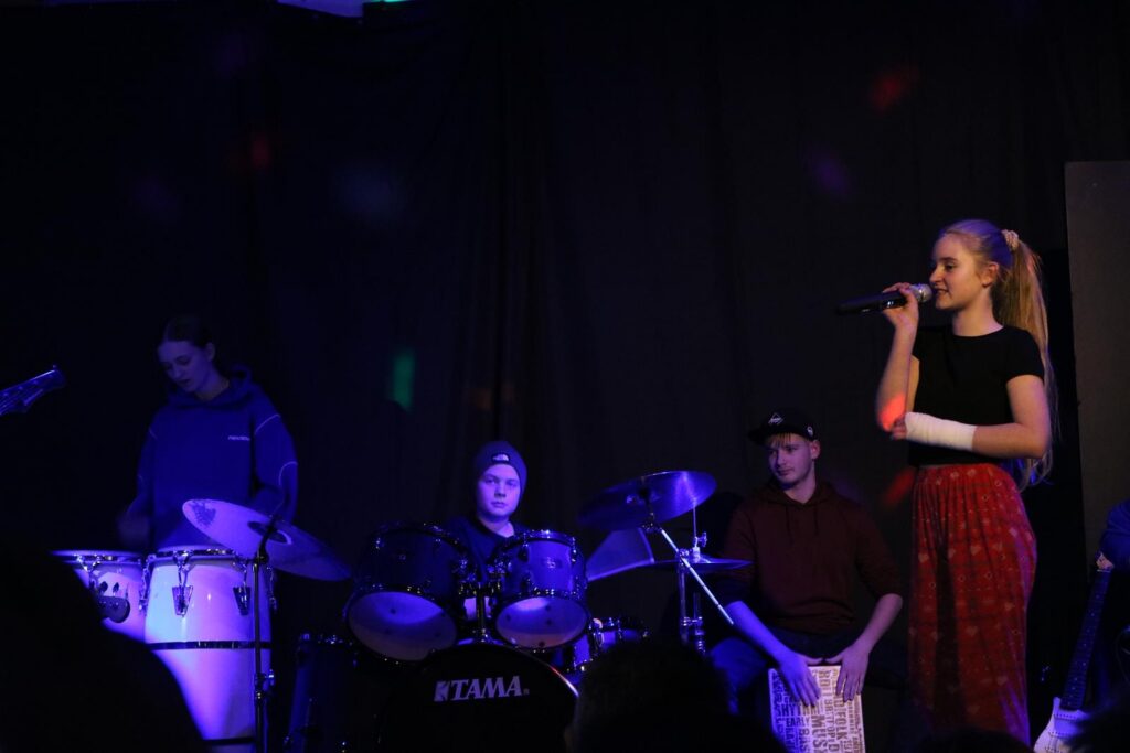 Schülerband tritt auf, Sängerin mit Mikrofon, Musiker an Schlagzeug und Percussion.