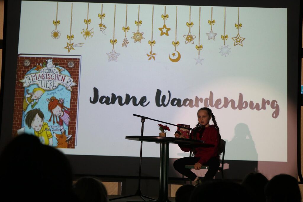 Ein Mädchen sitzt an einem Tisch und präsentiert ein Buch auf einer Bühne.