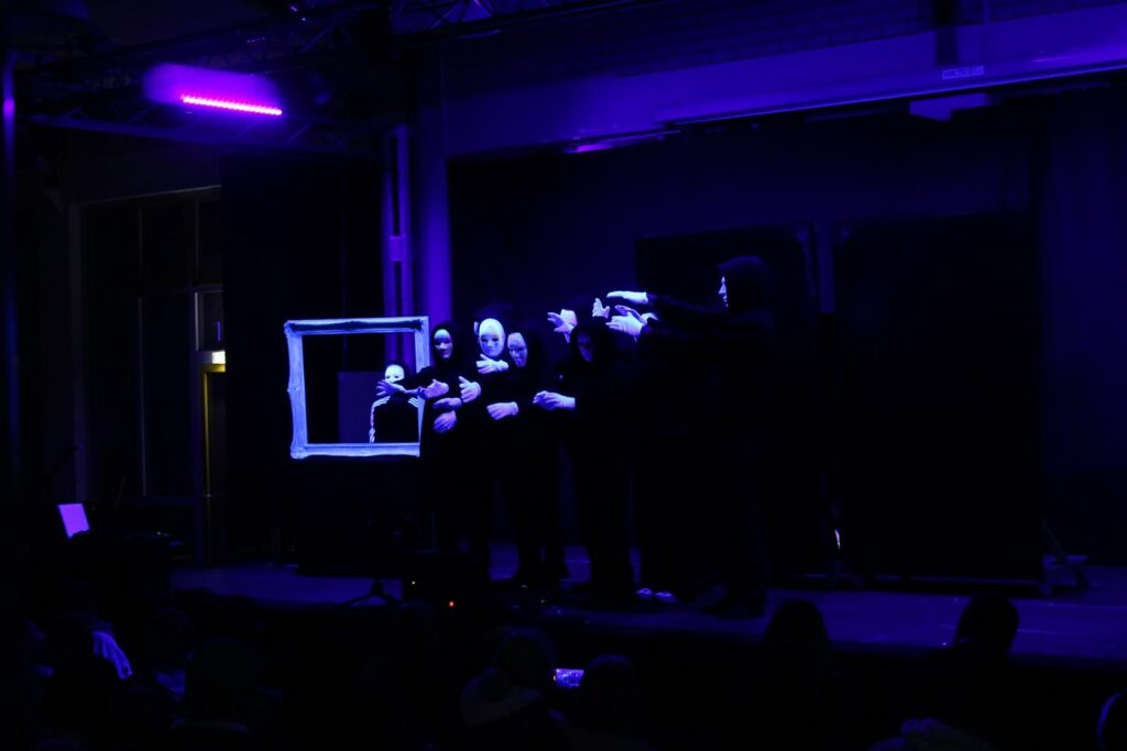 Theateraufführung mit mehreren Darstellern in schwarzer Kleidung und lila Beleuchtung.